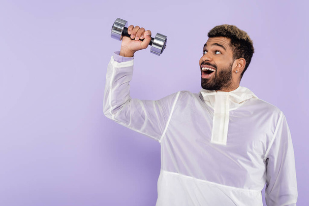 excited african american sportsman exercising with dumbbell isolated on purple  - 写真・画像