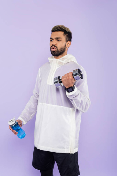 bearded african american sportsman exercising with dumbbell and holding sports bottle isolated on purple  - Фото, изображение