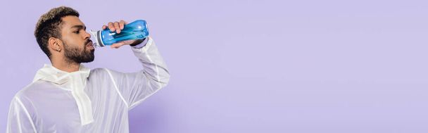 бородатый африканский американский спортсмен держит спортивную бутылку и пьет освежающую воду на фиолетовом, баннере - Фото, изображение