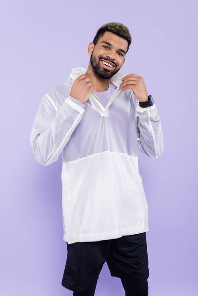 happy african american sportsman with smart watch adjusting collar isolated on purple  - Foto, afbeelding
