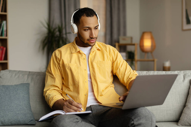 Vakava nuori musta mies kuulokkeissa, joilla on online-liiketapaaminen kannettavalla tietokoneella, kirjoittamalla tietoja kotona. Millennial Afrikkalainen Amerikkalainen mies opiskelija tekee muistiinpanoja aikana webinar, oppiminen etänä - Valokuva, kuva