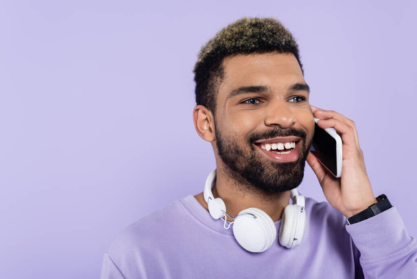 happy african american man in wireless headphones talking on smartphone isolated on purple  - Photo, image