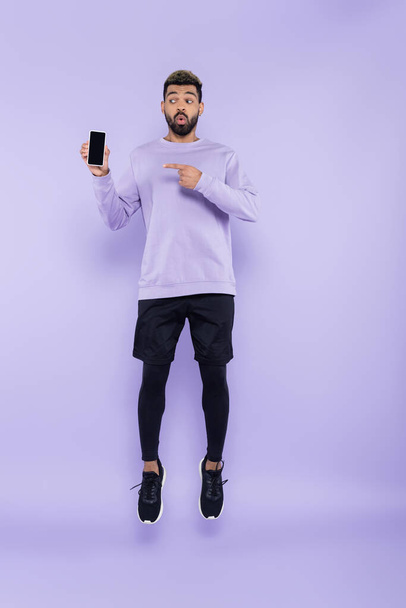 longitud completa de hombre afroamericano sorprendido apuntando al teléfono inteligente y levitando sobre púrpura - Foto, imagen