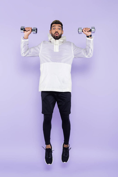 full length of shocked african american man in sportswear levitating and holding dumbbells on purple - Photo, Image