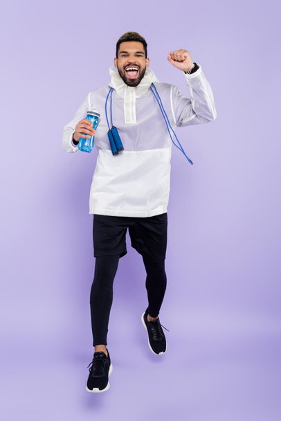full length of excited african american man in sportswear levitating and holding sports bottle on purple - Fotografie, Obrázek