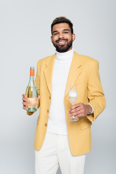 happy african american man in stylish outfit holding bottle and glass of champagne isolated on grey - Foto, Imagen
