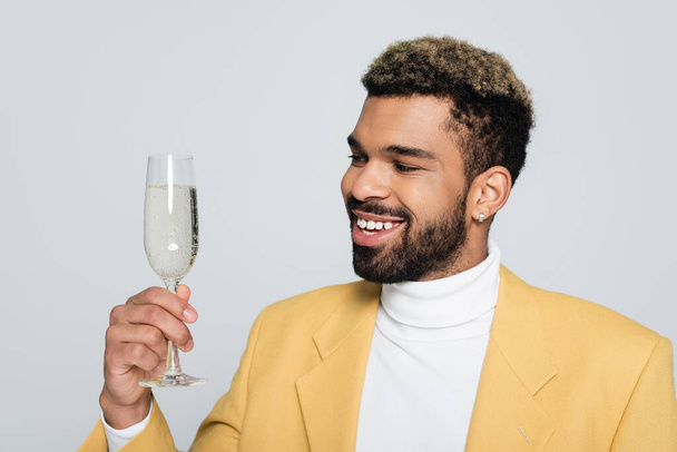 fröhliche afrikanisch-amerikanische Mann in stilvollem Outfit hält Glas Champagner isoliert auf grau  - Foto, Bild