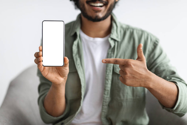 Cropped of bearded man in casual sitting in armchair, pointing at cell phone with white blank screen, recommending mobile application, white studio background, mockup, copy space - Fotó, kép