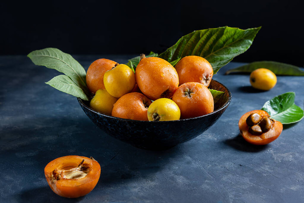 Keltainen ja oranssi Medlar hedelmiä lautasella lehtiä tummalla betonitaustalla, kaksi puolet hedelmistä pöydällä - Valokuva, kuva