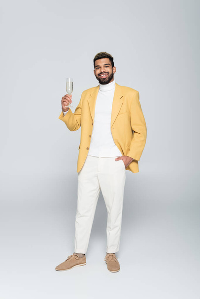 full length of cheerful african american man in stylish outfit holding glass of champagne and posing on grey  - Fotó, kép