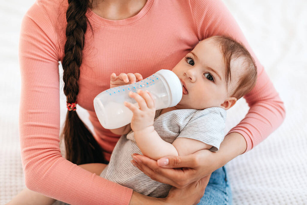 Closeup portrait of mother with baby on hands, little child holding bottle and drinking milk, looking at camera. Nutrition, love, care and family concept - Photo, Image
