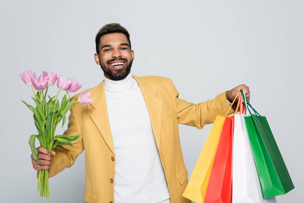 cheerful african american man in yellow blazer and polo neck holding pink tulips and shopping bags isolated on grey  - Foto, imagen