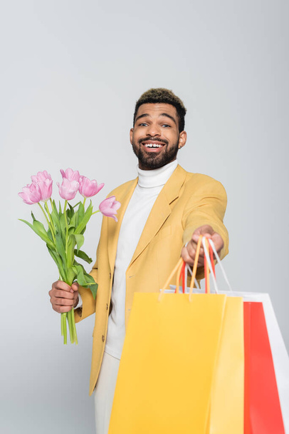smiling african american man in yellow blazer and polo neck holding pink tulips and shopping bags isolated on grey  - Foto, Imagen