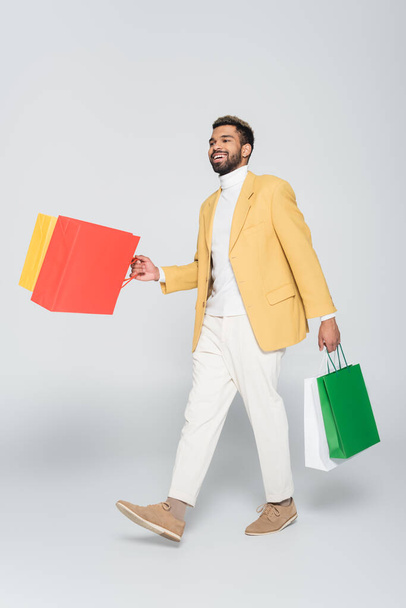 full length of cheerful african american man in yellow blazer holding shopping bags and walking on grey - Foto, Imagem
