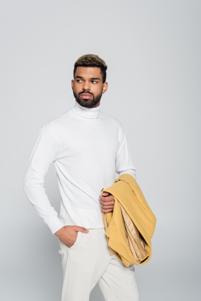 happy african american man with blue eyes in turtleneck holding blazer and posing with hand in pocket isolated on grey - Photo, Image