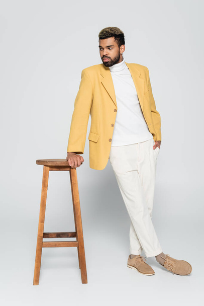 longitud completa de barbudo hombre afroamericano en traje elegante de pie con la mano en el bolsillo cerca de la silla alta de madera en gris - Foto, imagen