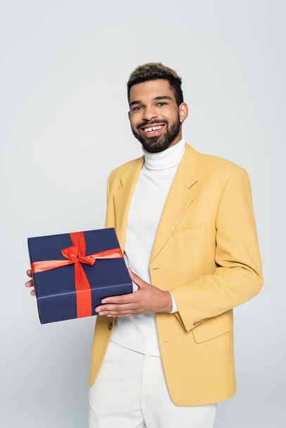 happy african american man in yellow stylish blazer holding wrapped present isolated on grey  - Fotografie, Obrázek