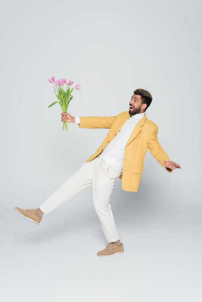 full length of exited african american man in stylish blazer holding bouquet of pink tulips on grey - Photo, image