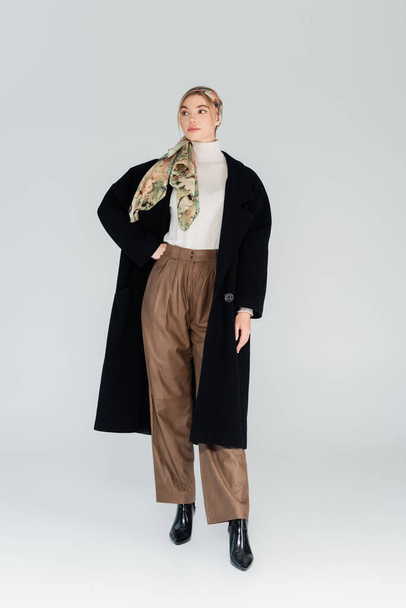 full length of woman in black coat and beige trousers posing with hand on waist on grey background - Fotoğraf, Görsel