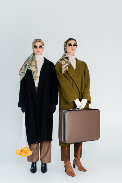 piena lunghezza di donne alla moda con valigia retrò e arance fresche in borsa a rete su sfondo grigio - Foto, immagini