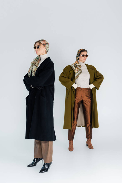 full length of women in coats and sunglasses posing with crossed arms and hands on hips on grey background - Фото, зображення