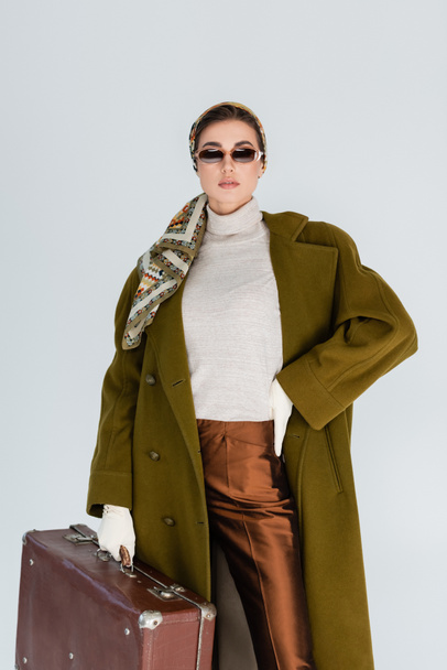 trendy woman in green coat and sunglasses posing with vintage suitcase and hand on waist isolated on grey - Photo, Image