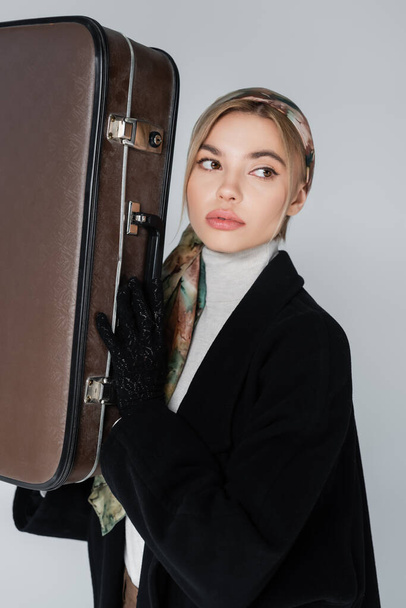 young woman in kerchief holding vintage suitcase isolated on grey - Foto, afbeelding