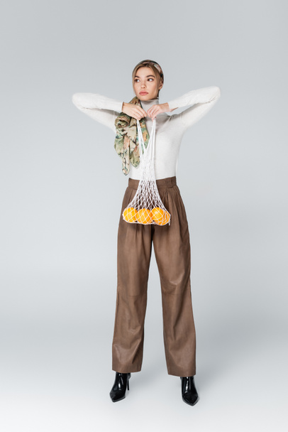 pełna długość kobiety w beżowych spodniach pozujących z pomarańczami w siatkowym worku na szarości  - Zdjęcie, obraz