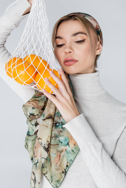 hezká žena v šátku pózuje s čerstvými pomeranči v síťované tašce izolované na šedé - Fotografie, Obrázek