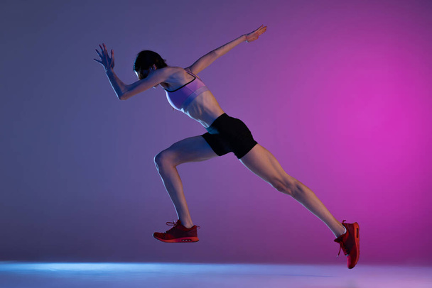 In motion. One young muscular girl, female runner or jogger training isolated on pink-blue background in neon light. Sport, track-and-field athletics, competition and active lifestyle concept. - 写真・画像