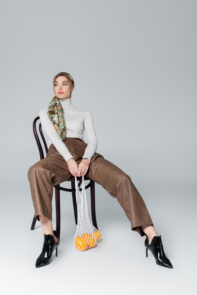 full length of woman in beige trousers sitting on chair with ripe oranges in net bag on grey background - Photo, Image