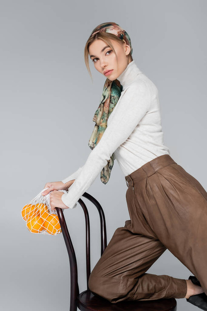stylish woman in turtleneck and kerchief posing with chair and net bag with oranges isolated on grey - Photo, Image