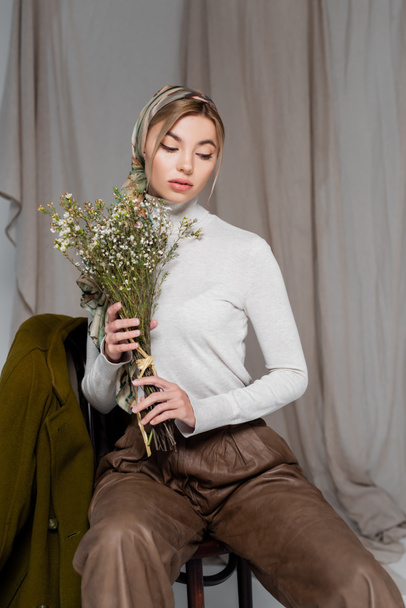 stylish woman in white turtleneck sitting with bouquet of gypsophila flowers on grey background - Foto, afbeelding