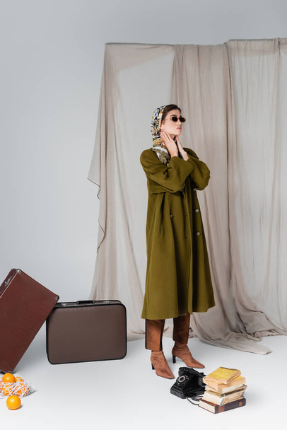 elegant woman in sunglasses standing by suitcases and vintage phone near books on grey draped background - Photo, Image