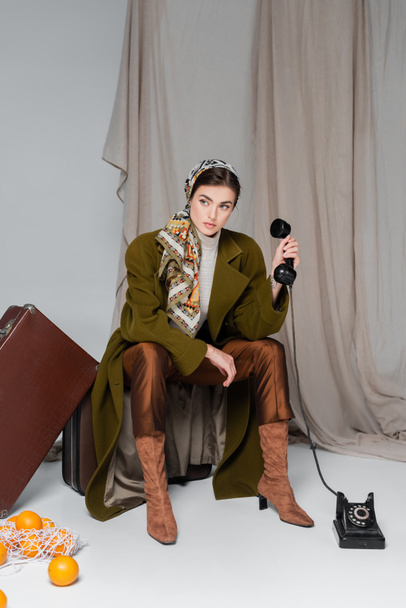 стильная женщина с ретро-телефоном, сидящая на чемодане рядом с апельсинами на сером фоне с тканью - Фото, изображение