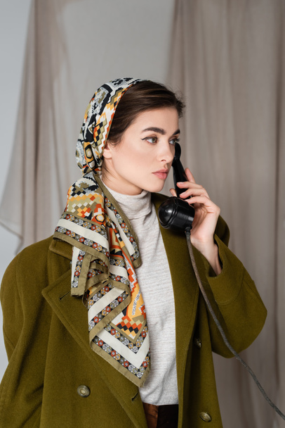 stylish woman in patterned headscarf calling on vintage telephone on grey background with drapery - Photo, Image