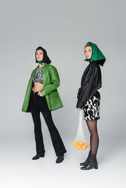 volle Länge der Frau, die mit Orangen in Netztasche neben trendigen Freund auf grauem Hintergrund steht - Foto, Bild
