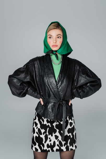 trendy woman in leather jacket and green kerchief posing with hands on waist isolated on grey - Photo, Image