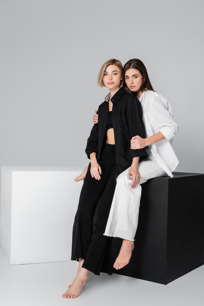brunette woman hugging friend wearing black clothes while sitting on cube on grey background - Photo, Image