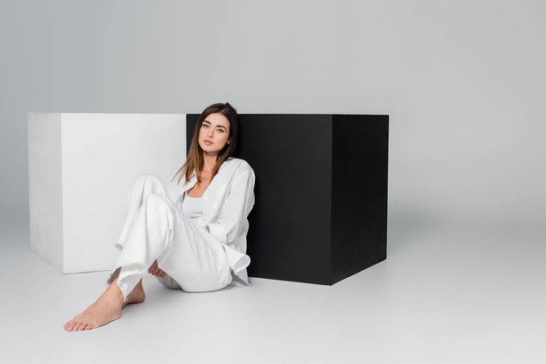 full length of barefoot woman sitting near black and white cubes on grey background - Photo, Image