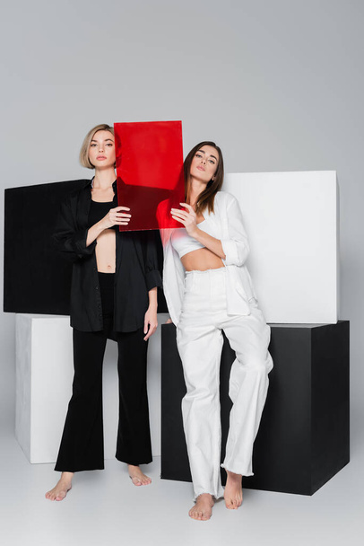 full length of barefoot women posing with red glass near black and white cubes on grey background - Fotó, kép