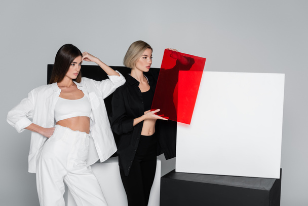 woman holding red glass while standing with friend near black and white cubes isolated on grey - Photo, Image