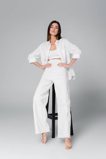 full length of barefoot woman in white clothing posing with hands on hips near black stool on grey background - Photo, Image