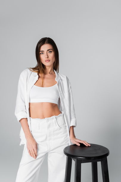 woman in white bra and shirt posing near black stool isolated on grey - Фото, изображение