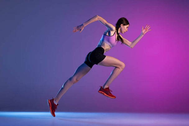 In action. One young muscular girl, female runner or jogger training isolated on pink-blue background in neon light. Sport, track-and-field athletics, competition and active lifestyle concept. - Fotó, kép