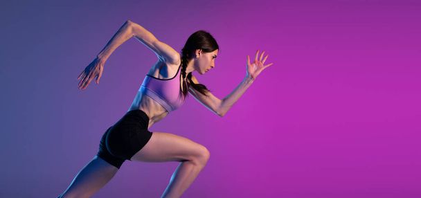 In motion. One young muscular girl, female runner or jogger training isolated on pink-blue background in neon light. Sport, track-and-field athletics, competition and active lifestyle concept. - Фото, зображення