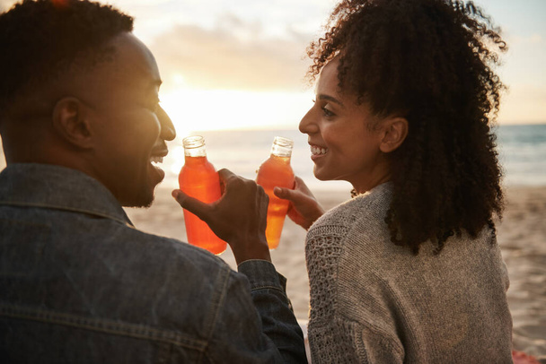 Rear view of a smiling young multiethnic couple drinking juice while sitting on a sandy beach at sunset - Foto, Imagen
