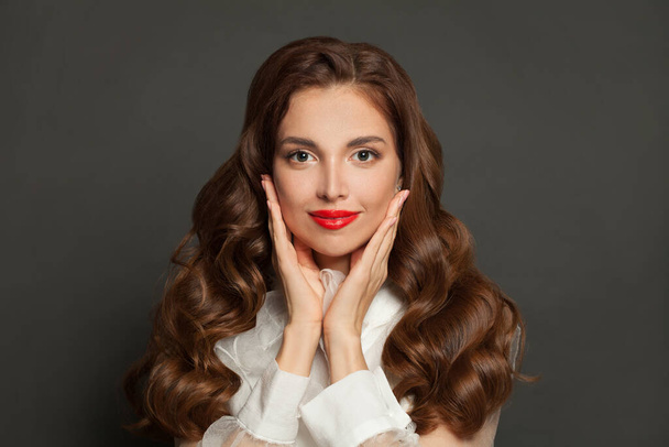 Belle femme aux longs cheveux ondulés tenant ses mains près de son visage. Brunette avec coiffure bouclée et lèvres rouges    - Photo, image