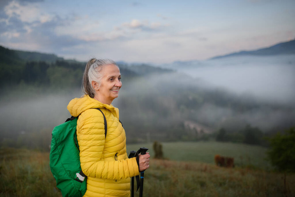 A senior woman hiking in nature on early morning with fog and mountains in background. - Фото, изображение