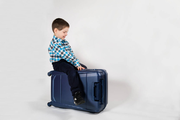 Um menino de 4 anos se senta em uma mala de viagem azul em um fundo branco com espaço para texto - Foto, Imagem